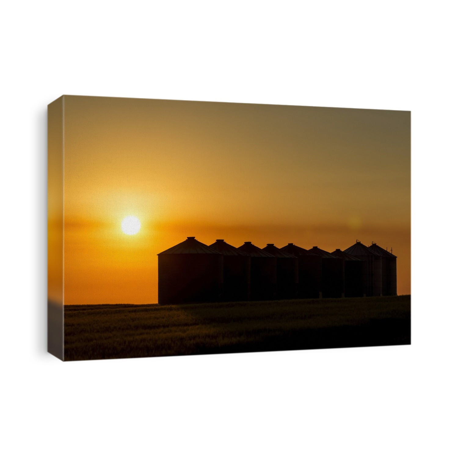 Silhouette of large metal grain bins at sunrise with an orange sun and an orange cast sky, East of Calgary; Alberta, Canada